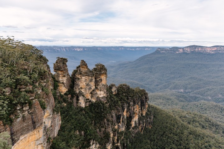 The Three Sisters rock formation in the Blue Mountains, Australia, with forested cliffs under a cloudy sky.