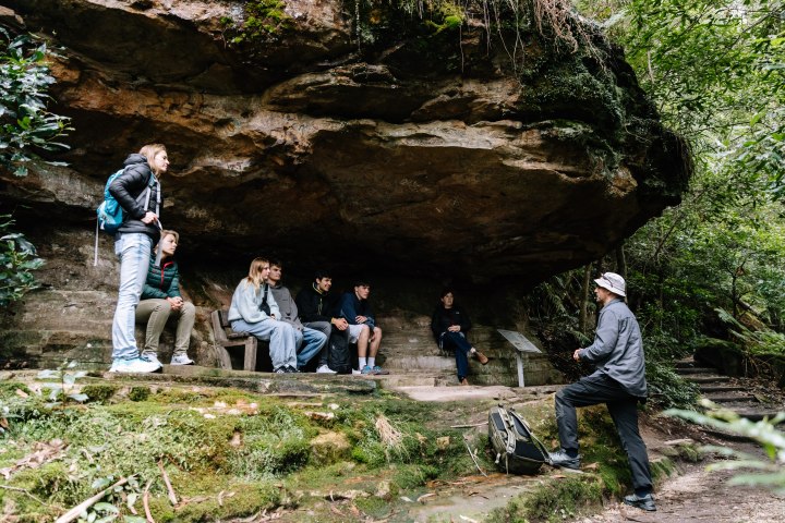 People seated under a rock overhang, one standing and one facing them, surrounded by greenery.