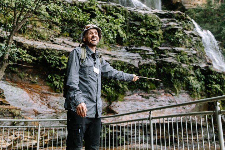 Man with backpack and hat smiles near a waterfall and railing.