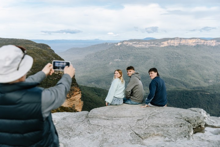 Three people sitting on a cliff while another person takes their photo with a phone.