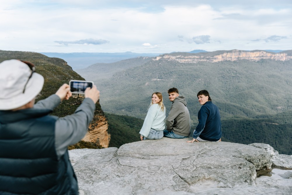 Three people sitting on a cliff while another person takes their photo with a phone.