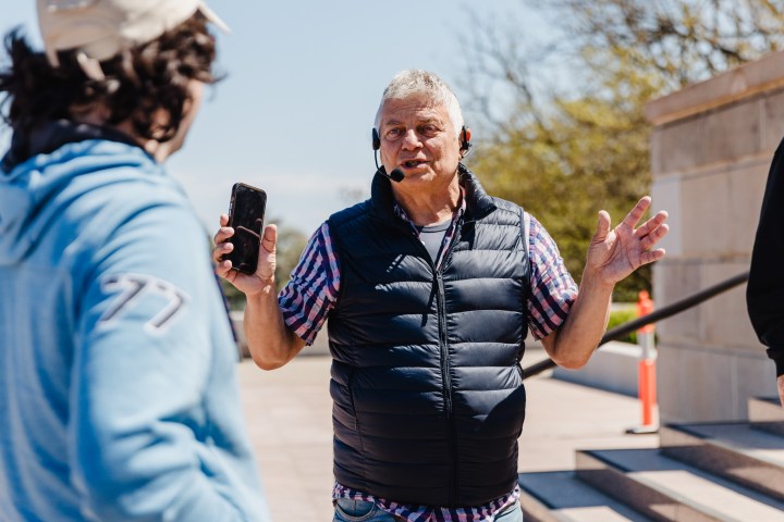 Man with headset talks and holds phone outside, others nearby, sunny day.