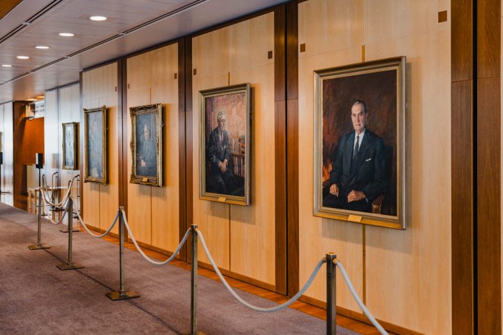 Hallway with framed portraits on wooden walls, separated by mannequins and velvet ropes.