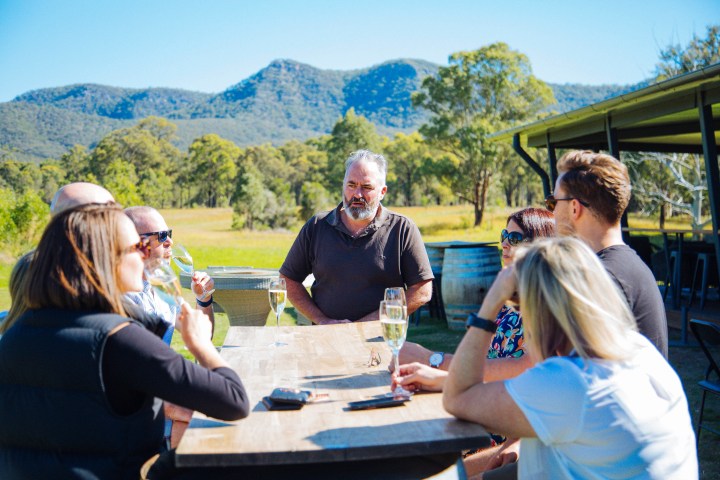 Group of people enjoying outdoor drinks by a wooden table with mountains in the background.