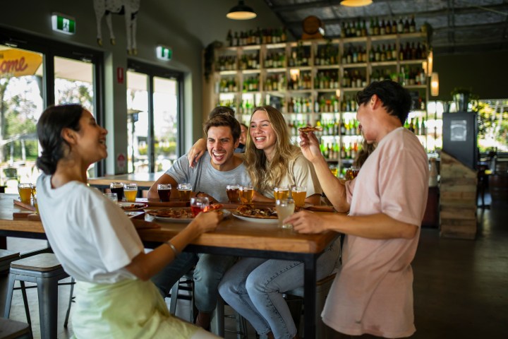 Four friends laughing and enjoying drinks and pizza at a bar with shelves of bottles in the background.