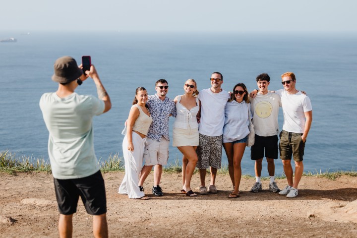 Group of seven people smiling on a seaside cliff while someone takes a photo, ocean in the background.