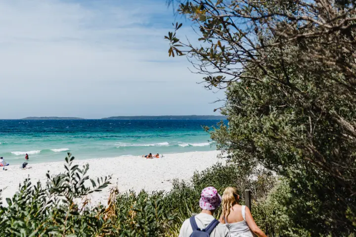 Two people walking down a sandy path to a beach with turquoise water and blue sky.