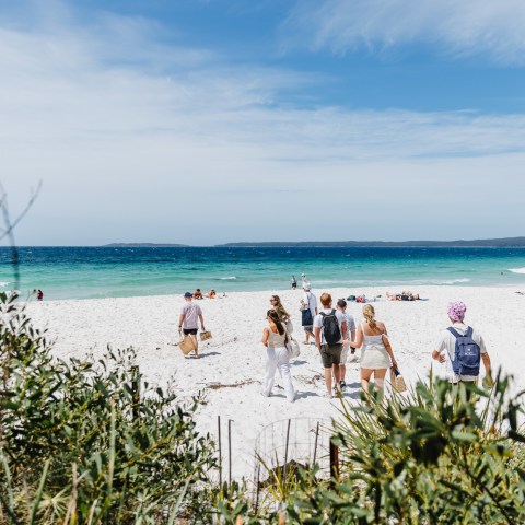 Group of people walking towards a sunny beach with white sand and turquoise water.