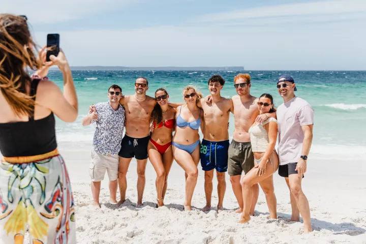 Group of friends posing for a photo on a sunny beach with waves in the background.