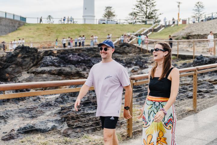 A man and woman in casual clothes walk by a rocky coastline with a lighthouse in the background.