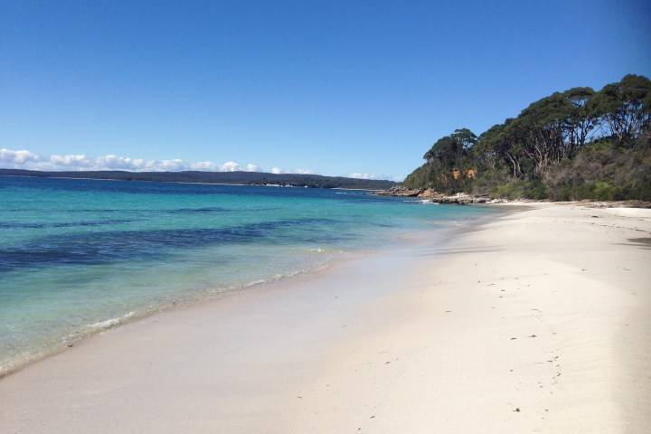 Sunny beach with white sand, clear blue water, and trees in the background.
