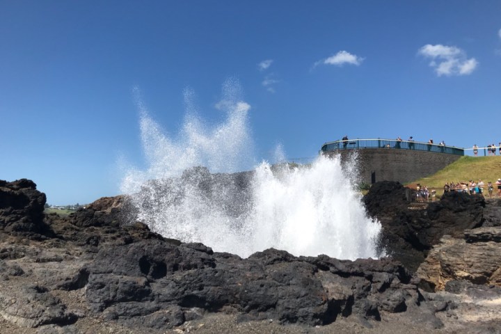 Water geyser bursts from rocky ground under a clear blue sky; people observe from a fenced platform.