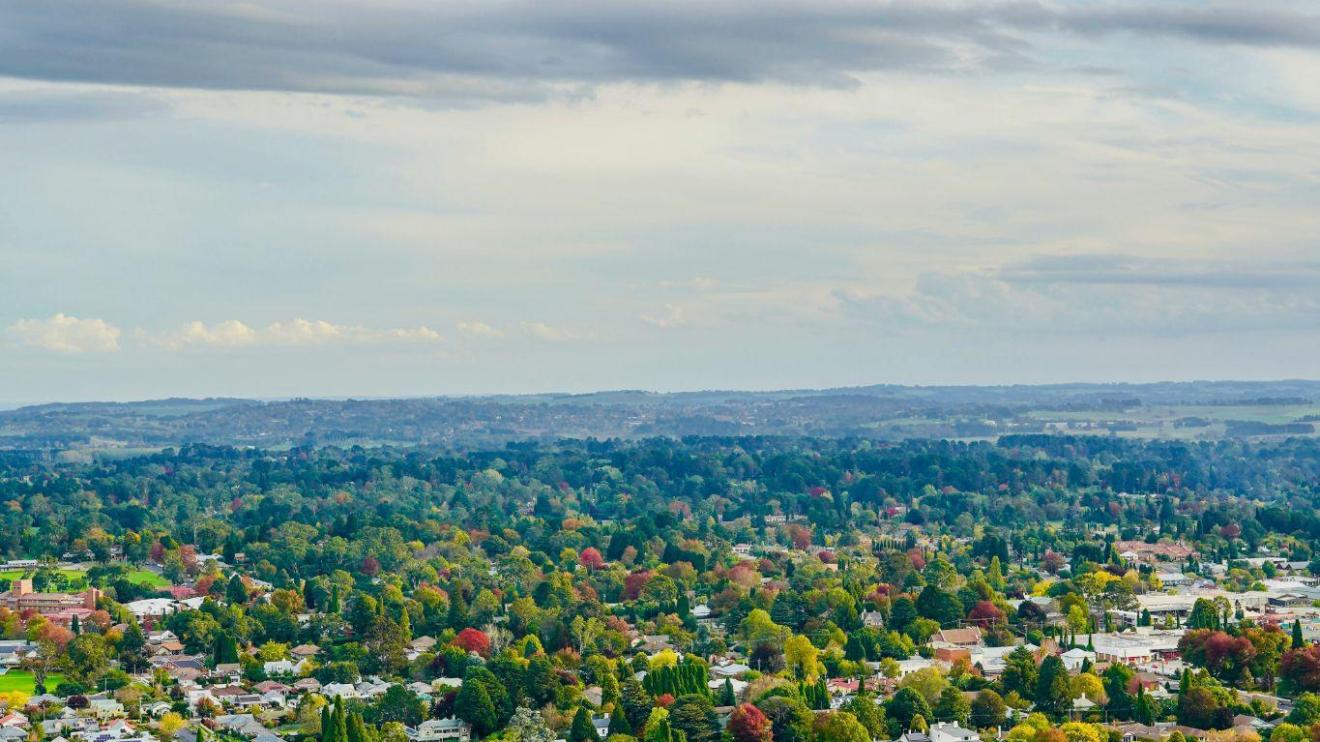 Aerial view of a town surrounded by vibrant green and autumnal trees under a cloudy blue sky.