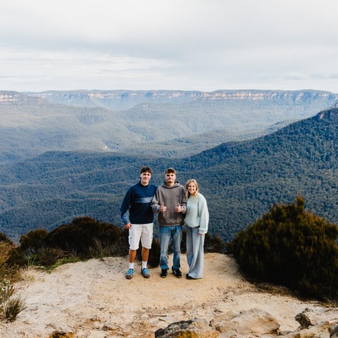Three people posing on a cliff with a view of mountains and forest in the background.
