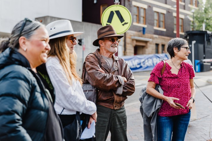 Four people in casual attire standing on a city street, one holding a jacket and another wearing a hat.