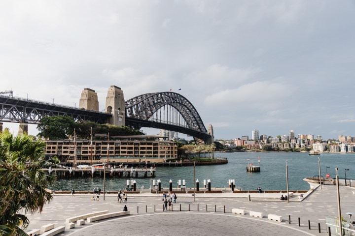 Sydney Harbour Bridge over water with city skyline and people walking.