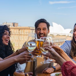 Group of friends toasting with drinks on a sunny rooftop, city skyline in the background.
