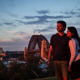 A couple poses together at sunset near a bridge with city skyline in the background.