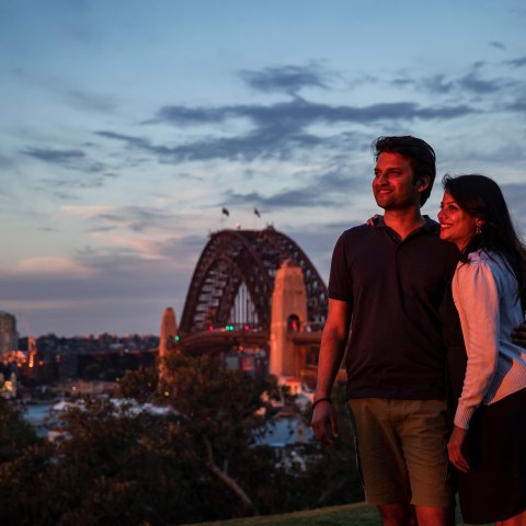 A couple poses together at sunset near a bridge with city skyline in the background.