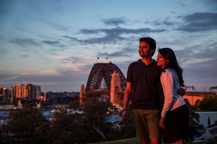 A couple poses together at sunset near a bridge with city skyline in the background.
