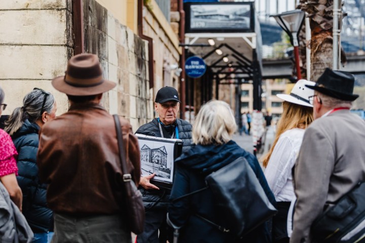 A tour guide shows an image to a group on a street near a bridge.