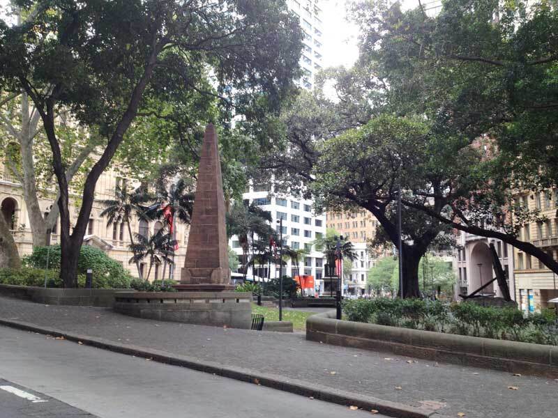 City park with trees, an obelisk, and surrounding buildings.