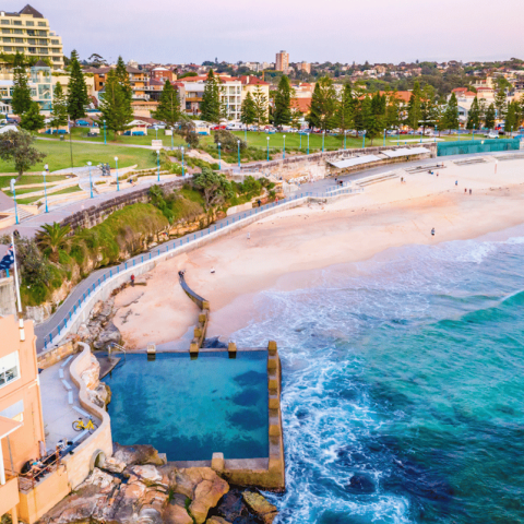 Coastal view of a beach with a pool, buildings, and people along the shore.