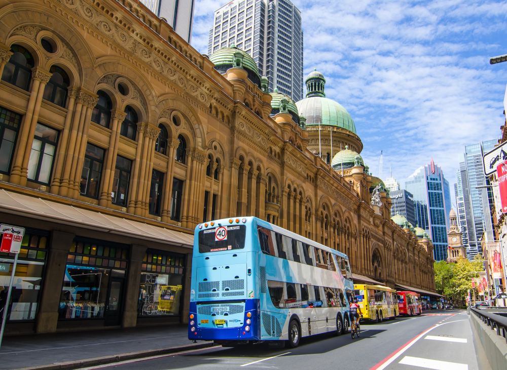 Double-decker bus on city street with historic building and skyscrapers.